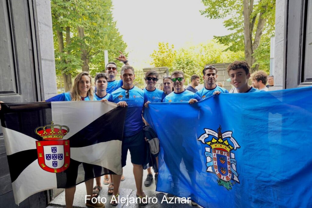 Group of people in blue shirts posing outdoors while holding a Portugal flag (left) and a blue flag with a crest (right).