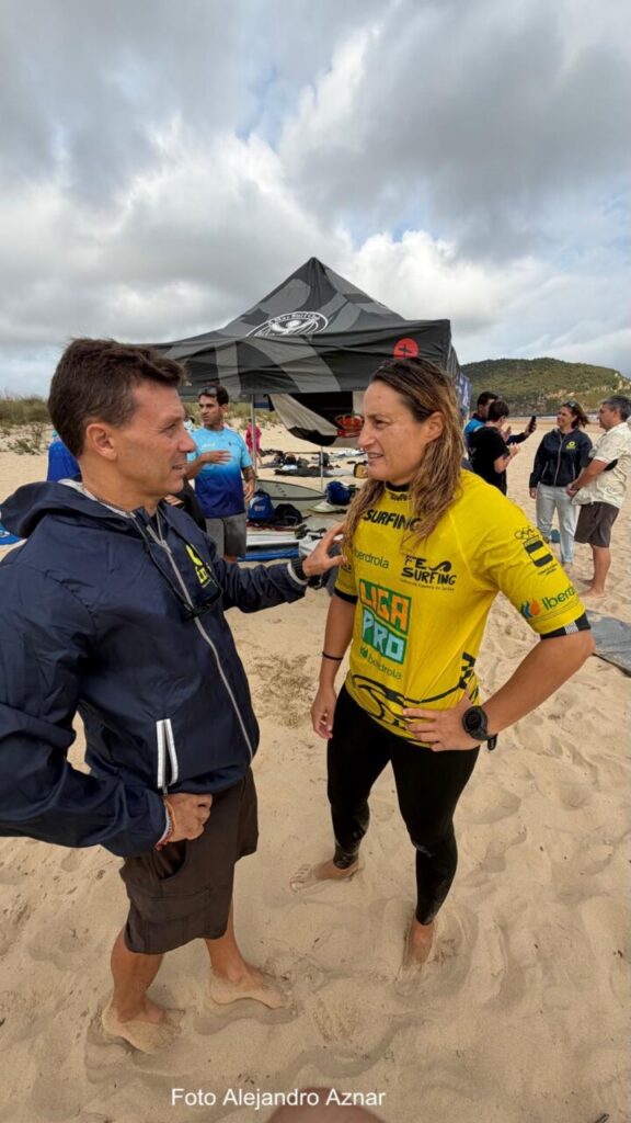 Two men on a sandy beach talk near a dark canopy at a surfing event; the woman in a yellow sponsor jersey stands nearby.
