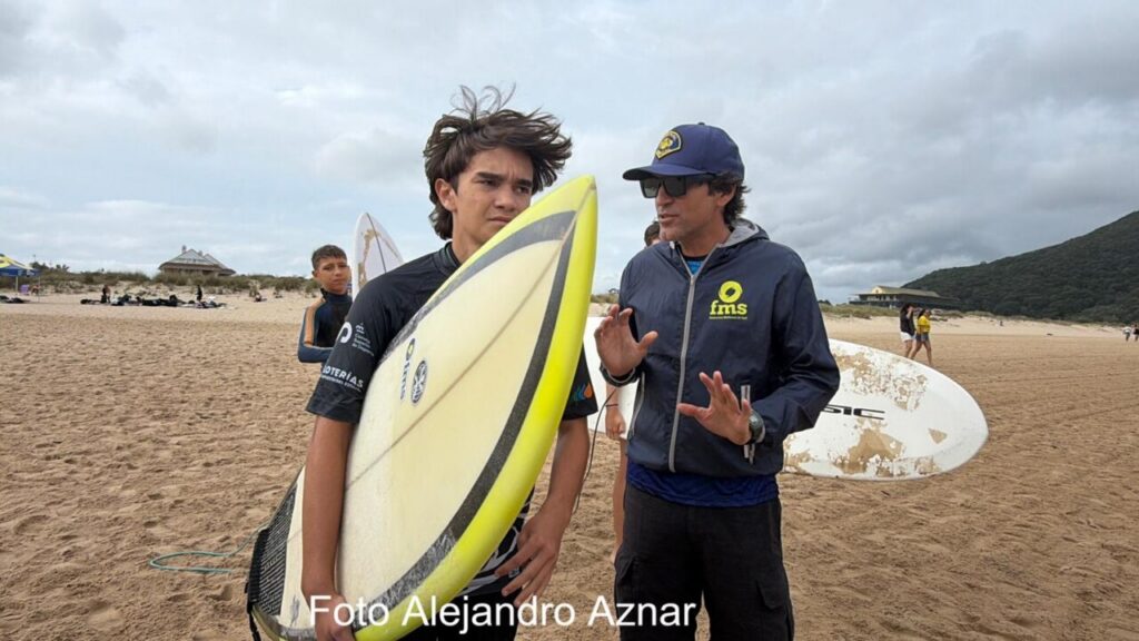 Young surfer holds a yellow surfboard on the sandy beach while a coach in a blue jacket talks to him; another board rests in the background.