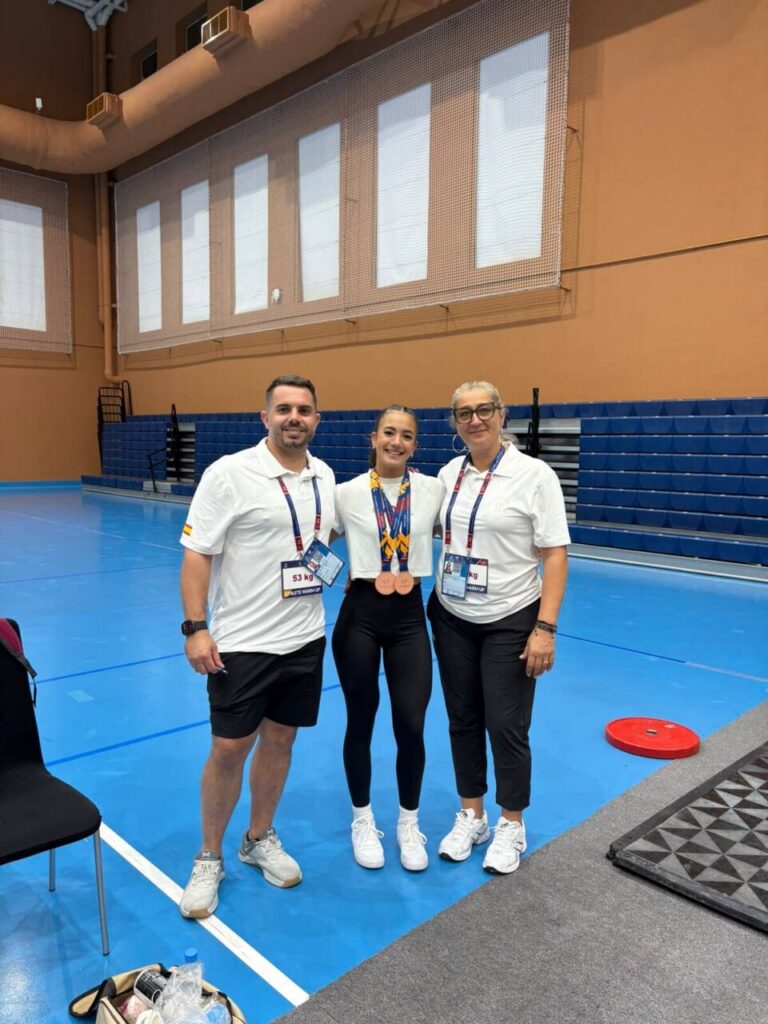 Young female athlete with two bronze medals posing with her two coaches in a gymnasium on a blue court, all wearing white shirts and lanyards.