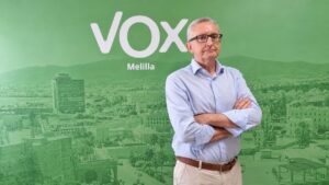 Man with arms crossed in front of a green Vox Melilla backdrop, wearing a light blue shirt and beige pants.