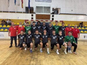 Indoor gym team photo: handball squad in black jerseys with numbers, coaches in red shirts, yellow bleachers in background.