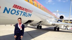 Flight attendant in uniform standing on tarmac beside an Iberia Regional Nostrum airplane with yellow stripe on the tail.