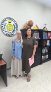 Two women posing together in an office, one in a beige hijab and blue top, the other in a dark green dress, standing in front of a branding plaque and shelves with books and trophies on the wall behind them.