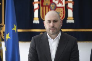 Man in a dark suit and white shirt seated indoors, looking ahead, with a European Union flag and a heraldic banner in the background.