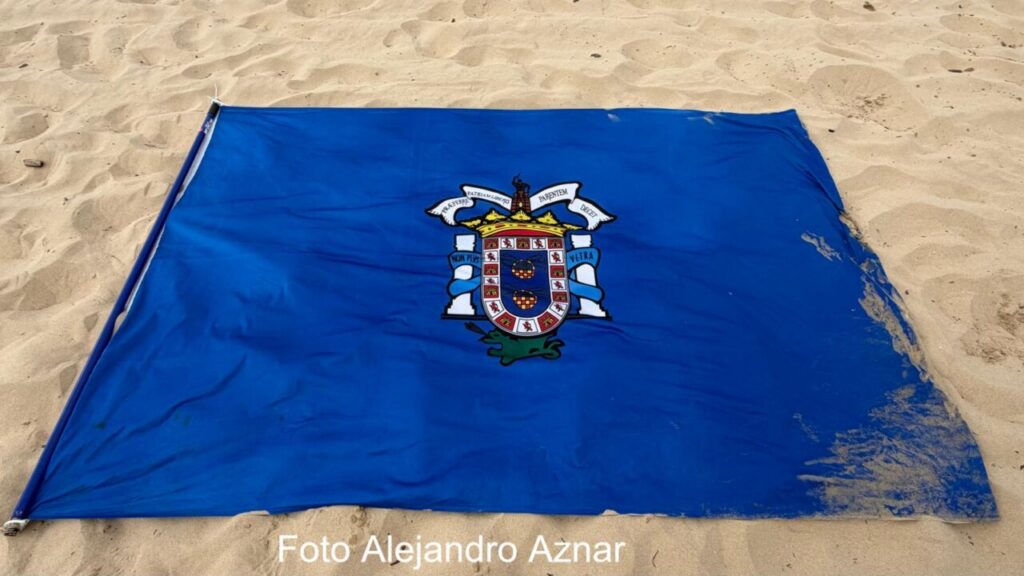 Blue flag with a coat‑of‑arms crest spread on a sandy beach, a flagpole lies at the left edge.