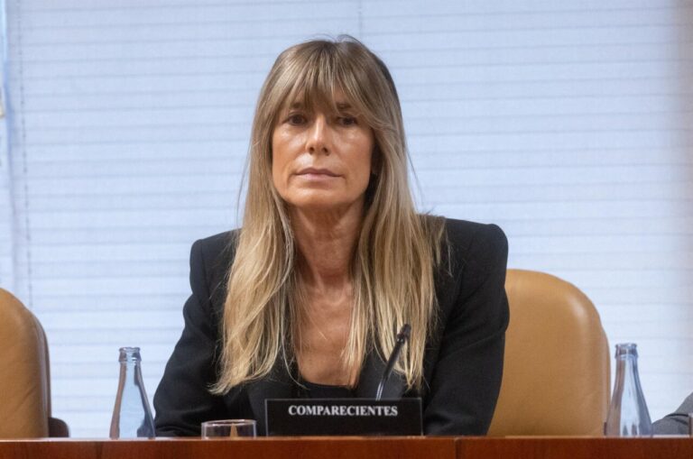 Professional woman in a black blazer seated at a conference table, facing forward with a microphone in front of her.