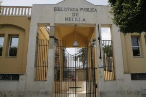 Entrance to the Melilla Public Library: cream stone gate with 'Biblioteca Publica de Melilla' and iron gates leading inside.