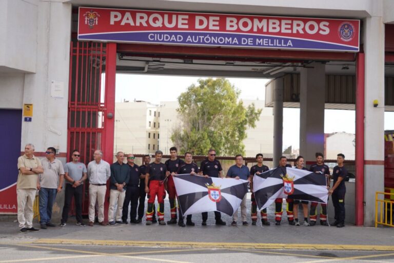 Group of firefighters standing in front of a fire station entrance holding black-and-white flags with a crest in Melilla