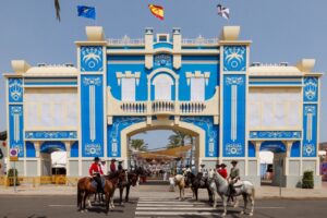 Blue ornate arch gateway with white decorative patterns, riders on horses in front at a crosswalk during a festival.