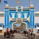 Blue ornate arch gateway with white decorative patterns, riders on horses in front at a crosswalk during a festival.