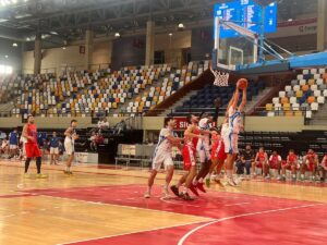 Basketball players in white and red uniforms contest a jump shot near the hoop in a crowded indoor arena with colorful empty seats in the background.
