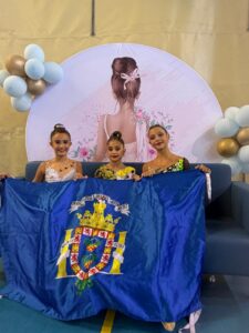 Three young dancers pose behind a blue flag with a royal crest, in front of a pastel backdrop with balloons and artwork.