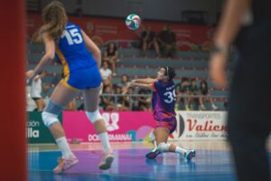 Action shot of a female volleyball player in a purple uniform (number 39) diving to pass the ball on an indoor court as a blue-uniform opponent approaches, with spectators in the background.