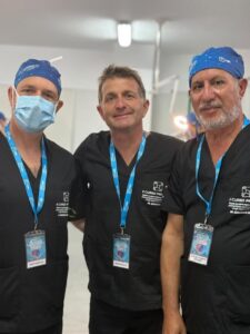 Three healthcare professionals in black scrubs with blue lanyards pose for a group photo in a clinical setting, all wearing ID badges and caps/facemasks on the left.