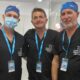 Three healthcare professionals in black scrubs with blue lanyards pose for a group photo in a clinical setting, all wearing ID badges and caps/facemasks on the left.