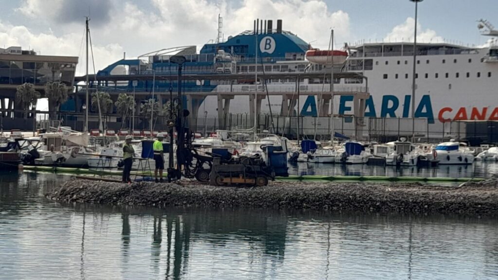 Marina scene with a large white ferry in the background and workers in reflective vests on a rocky breakwater near small boats.
