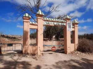 Entrance archway with pink pillars and green-topped capitals, a barred gate, and a no-entry sign in a dusty desert landscape; text on the sign reads 'Villa' and the area appears arid with a dirt road beyond the gate.