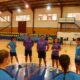 Group of players and coaches form a circle on an indoor gym floor, listening to a speaker at center court.