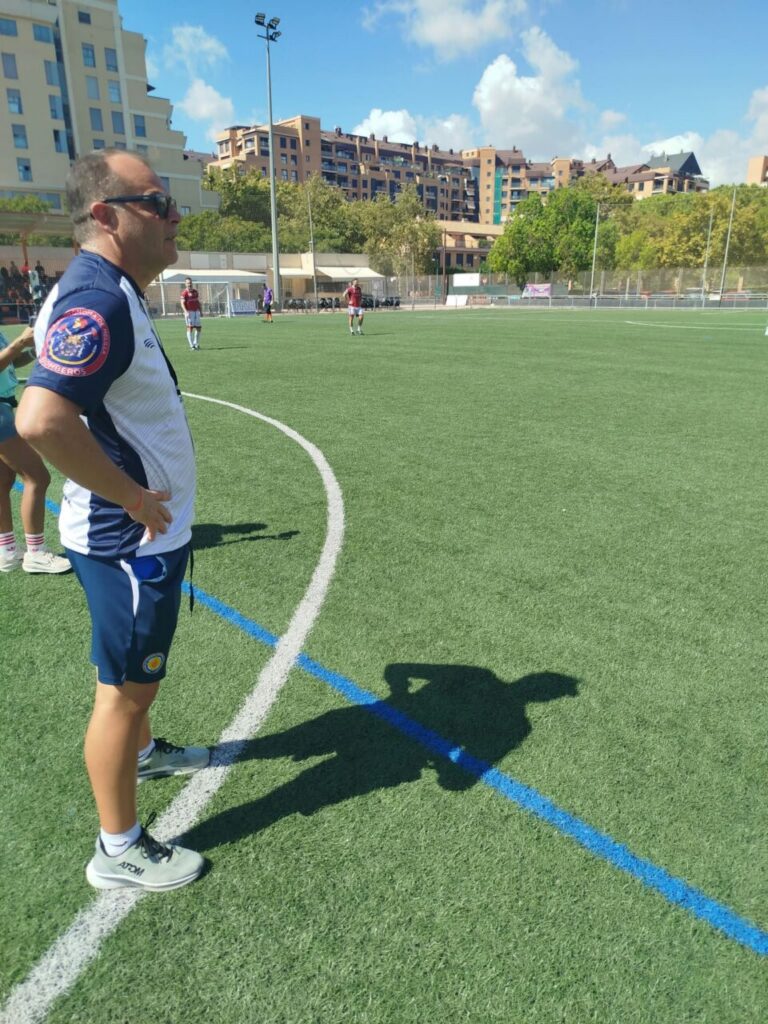Male coach standing on the sideline of a sunny soccer field, hands on hips, wearing a white and blue kit with a team patch on the sleeve, with players and apartment buildings in the background.