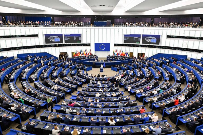 European Parliament chamber with circular blue desks and many delegates during a session, EU flag at center.