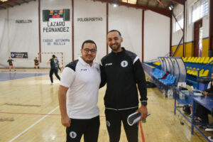 Two smiling men in sportswear pose inside a handball gym, with a scoreboard and blue-yellow seats in the background.