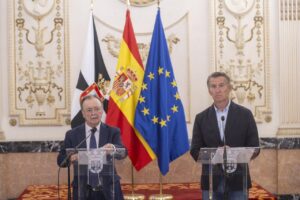 Two men stand at podiums giving a press conference, with the Spanish, EU, and another flag in the background in a formal ornate room.