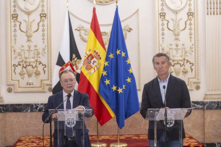 Two men stand at podiums giving a press conference, with the Spanish, EU, and another flag in the background in a formal ornate room.