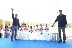 Two men on a blue stage at an outdoor event; one waves, the other points, as a crowd applauds behind them.