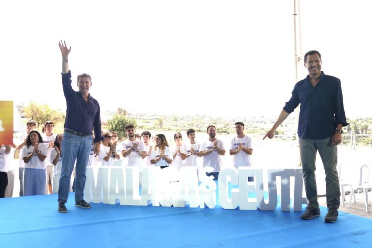 Two men on a blue stage at an outdoor event; one waves, the other points, as a crowd applauds behind them.