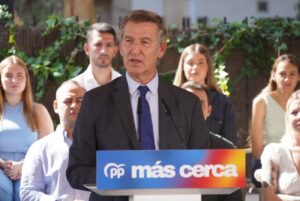 Man in a dark suit and blue tie speaks at a podium with a PP 'más cerca' banner, outdoors with supporters in the background.