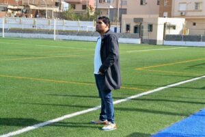 Man standing on a green artificial turf field with white lines, wearing a dark coat, white shirt, jeans, and sneakers, looking to the right.