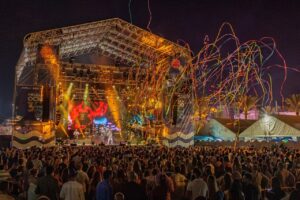 Crowd at a nighttime outdoor concert with a large stage, colorful lights, and streamers flying above the audience.