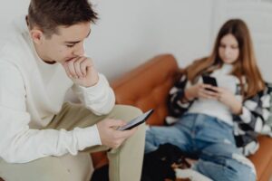 Young man and woman sit on a beige sofa absorbed in their smartphones, with a small dog at their feet.
