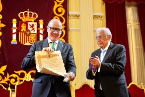 Two men in formal suits on a stage, one presenting a framed certificate and a small box while the other holds a blue ribbon medal.
