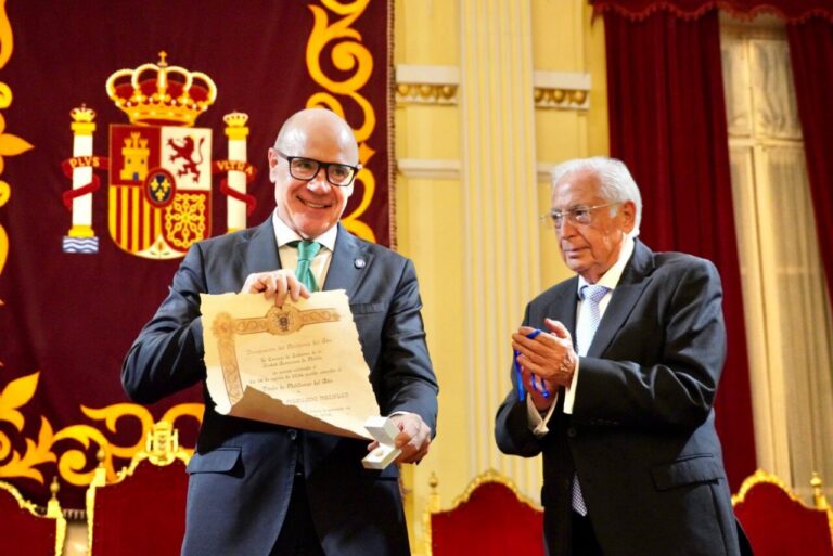 Two men in formal suits on a stage, one presenting a framed certificate and a small box while the other holds a blue ribbon medal.