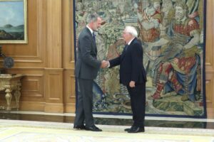 Two men in suits shake hands in a formal room with a large medieval-themed tapestry behind them and wooden paneling nearby