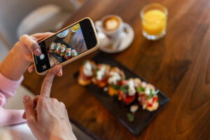 Person photographing a colorful sushi platter with a smartphone on a wooden table, with coffee and orange juice in the background.