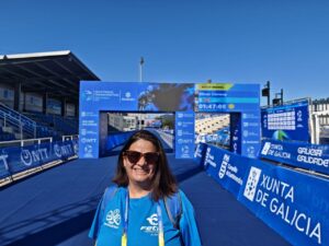 Smiling woman with sunglasses at a blue finish-line arch of a race, wearing a blue shirt and lanyard, event banners around.