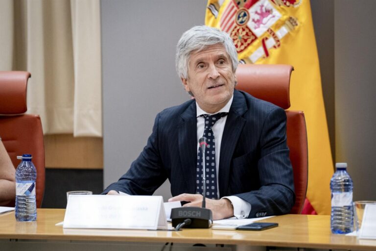 Middle-aged man in a dark pinstripe suit speaking at a conference table, with a microphone and nameplate in front, and a yellow flag in the background.