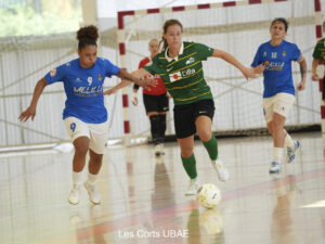 Futsal match: a player in a green jersey advances with the ball while two players in blue chase on an indoor court.