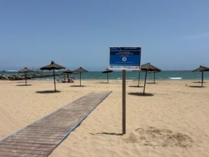 Beach scene with a wooden boardwalk leading to a blue sign on a post, thatched umbrellas, and people by the sea.