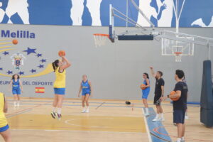 Female player in yellow shoots a jump shot while teammates watch in an indoor basketball court marked with Melilla signage.