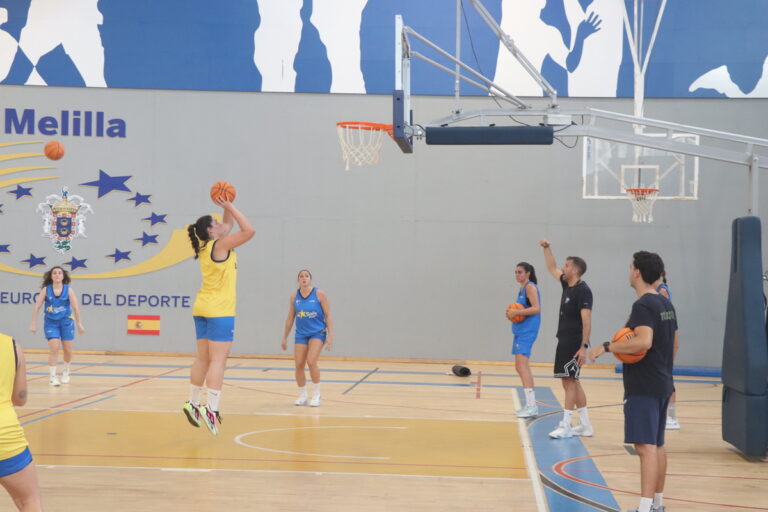 Female player in yellow shoots a jump shot while teammates watch in an indoor basketball court marked with Melilla signage.