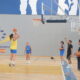 Female player in yellow shoots a jump shot while teammates watch in an indoor basketball court marked with Melilla signage.