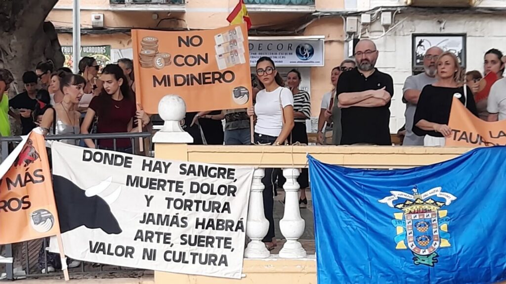 Crowd of protesters behind a railing with banners opposing funding and torture; a blue crest flag is on the right.