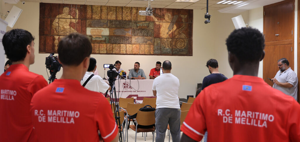 Press conference with panelists at a table facing reporters and cameras, in a room with a mural on the wall.