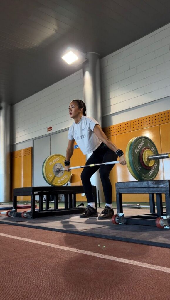 A female weightlifter in a white t‑shirt and black leggings lifts a loaded barbell from a platform in a gym, preparing for a clean or deadlift move.