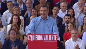 Man in a light blue shirt speaks at a red podium labeled 'IZQUIERDA VALIENTE' as a diverse audience claps behind him.