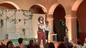 Actor in period costume gesturing on a small stage, another performer by a table; audience watches intently.